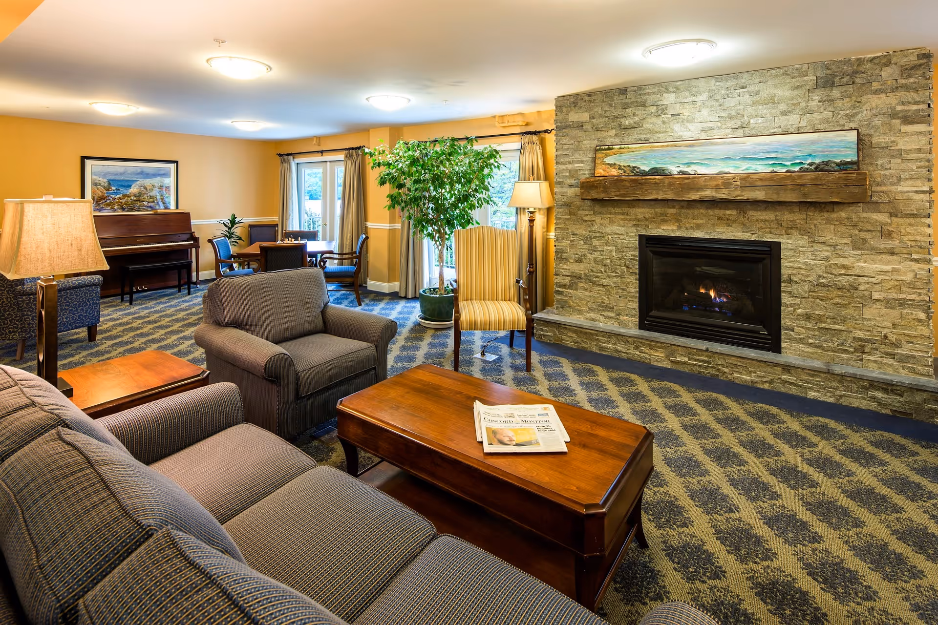 A cozy living room in an assisted living facility featuring a stone fireplace with a wooden mantel and a landscape painting above it. The room has a patterned carpet, a gray sofa, an armchair, a wooden coffee table with a newspaper on it, a striped armchair, a floor lamp, and a piano in the background near a table with chairs. Large windows with curtains allow natural light into the room.