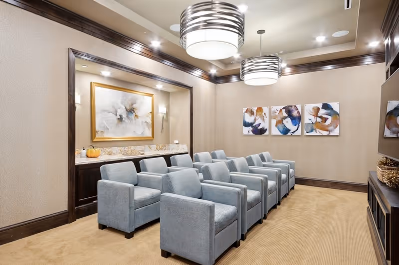 A small theater-style room with two rows of six light blue upholstered armchairs facing a wall-mounted TV. The room has beige walls with dark wood trim, abstract artwork on the walls, two modern circular ceiling lights, and a built-in counter with a decorative pumpkin and framed art above it.
