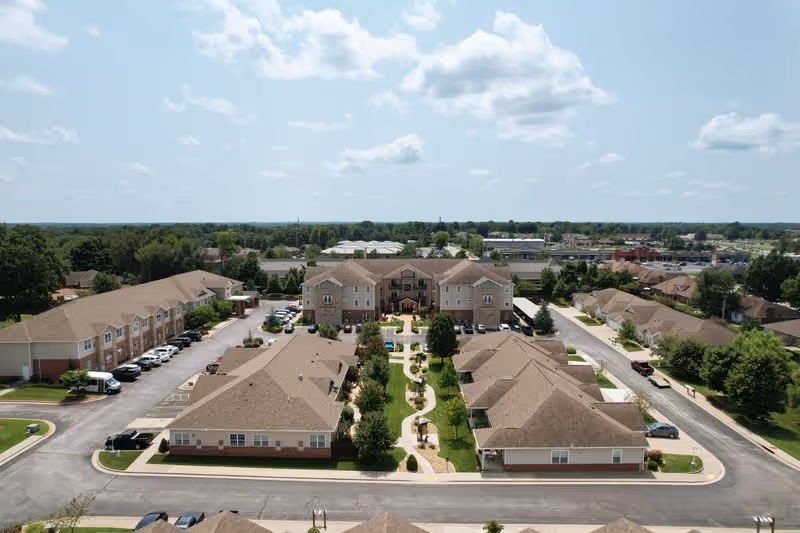 Aerial view of The Fremont Senior Living facility showing multiple beige buildings with brown roofs arranged around a landscaped courtyard with walking paths and greenery. Cars are parked along the driveways surrounding the buildings under a partly cloudy sky.