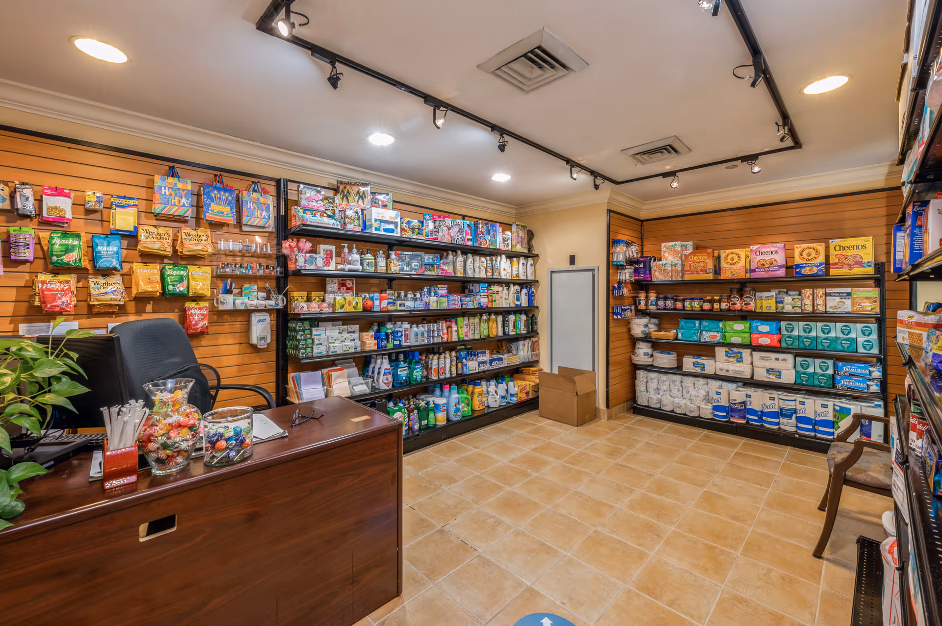 Interior view of a small convenience store or shop area within a senior living facility. Shelves are stocked with various items including snacks, cereals, personal care products, and household supplies. A wooden counter with a chair and some candy jars is visible in the foreground. The room has tiled flooring and track lighting on the ceiling.