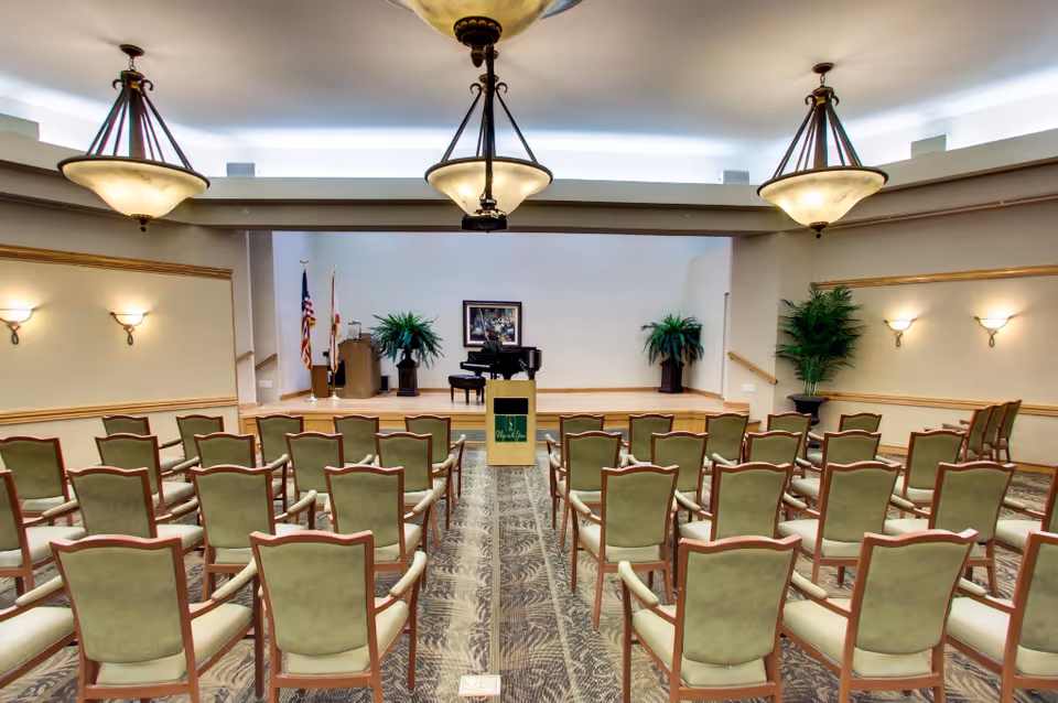 A medium-sized auditorium or meeting room with rows of green cushioned chairs facing a small stage. The stage has a podium with the Village on the Green logo, a grand piano, two large potted plants, and flags in the corner. The room is well-lit with three large ceiling light fixtures and wall sconces.