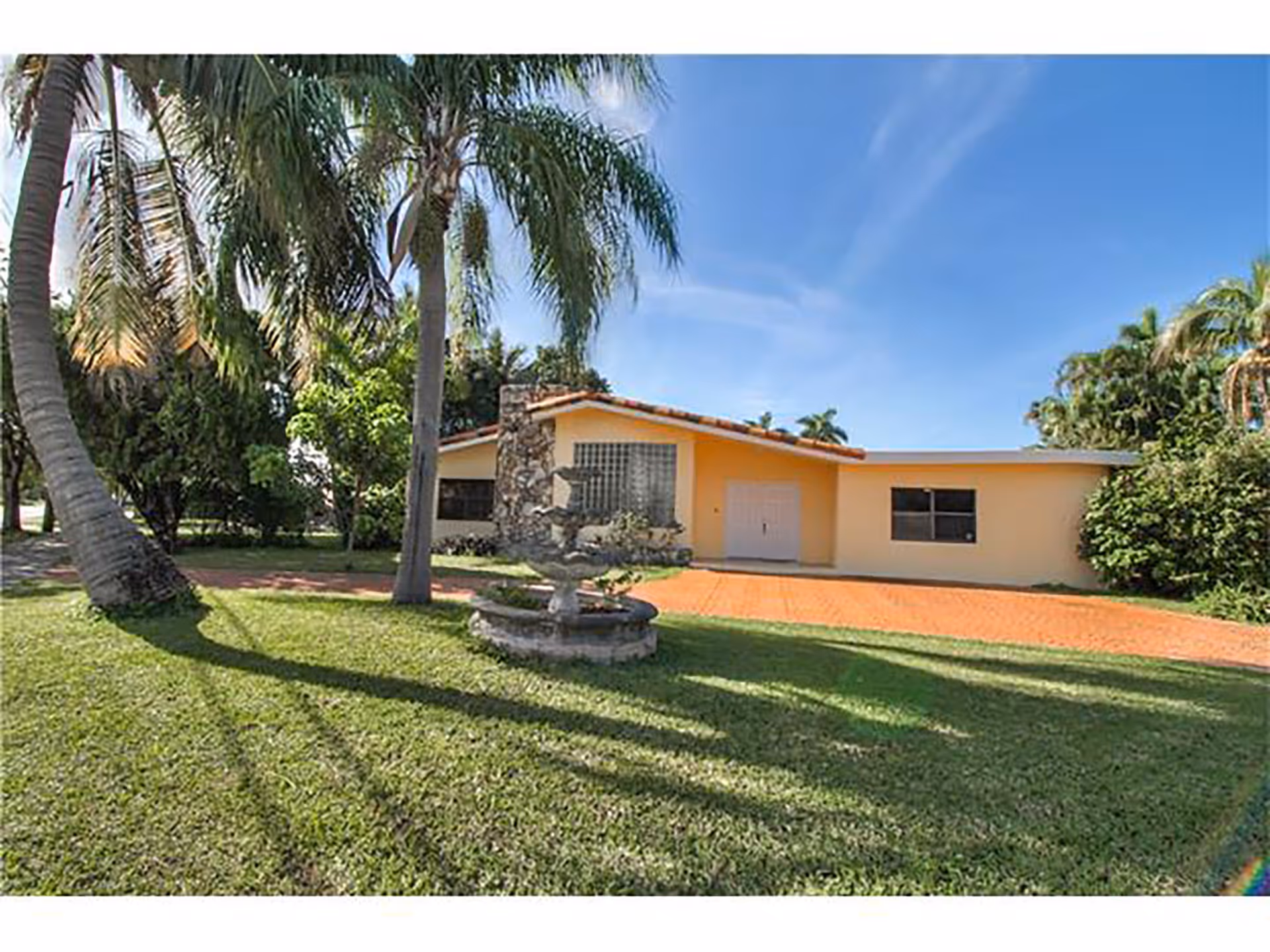 Single-story yellow house with a tiled driveway, palm trees and a small fountain on a grassy front lawn under a blue sky.
