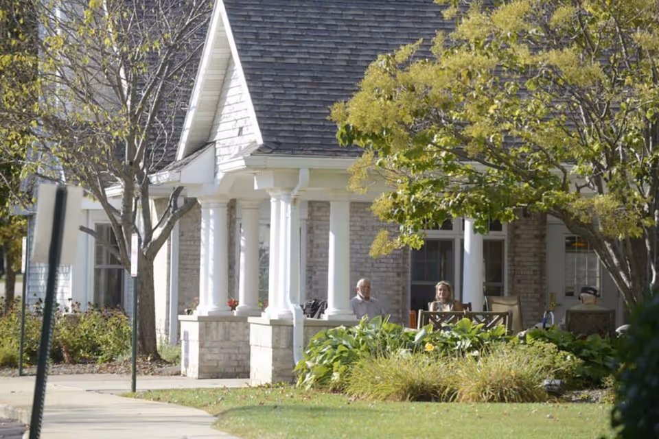 Outdoor patio area of a senior living facility with a few elderly people sitting on chairs under a covered porch surrounded by trees and greenery.