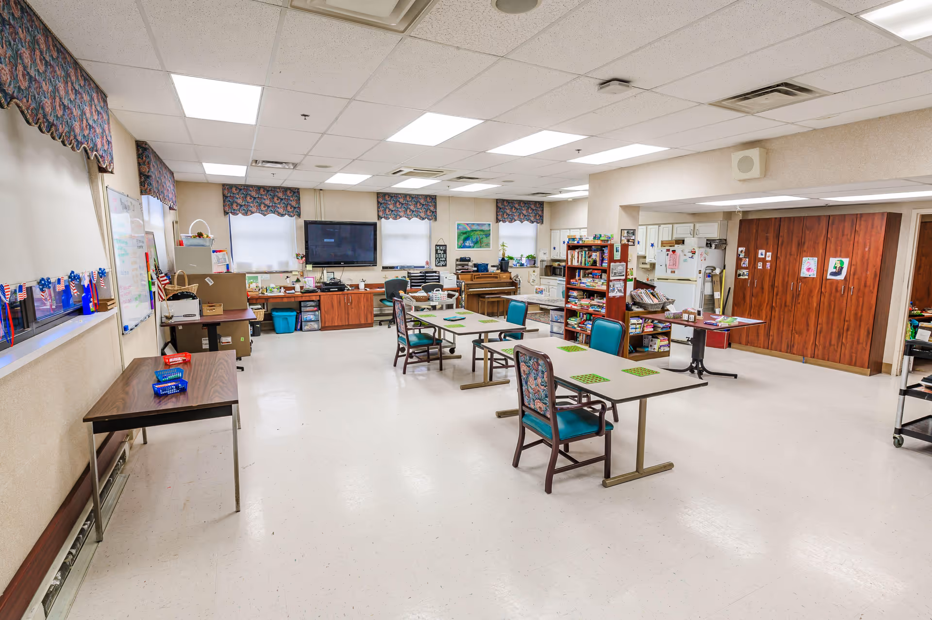 Large multipurpose activity room with tables, chairs, a TV, shelving and a kitchenette area.