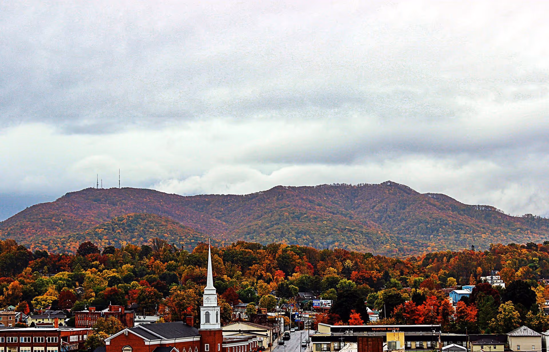 Autumnal small town with a church steeple in the foreground and tree-covered hills under a cloudy sky.