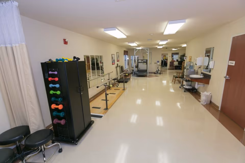 A rehabilitation room with exercise equipment including a rack of colorful dumbbells, parallel bars for walking practice, a treadmill, and other fitness machines. The room has bright overhead lighting, beige walls, and a clean, polished floor. There is a sink and hand sanitizer station on the right wall, and a door labeled 'Storage'.