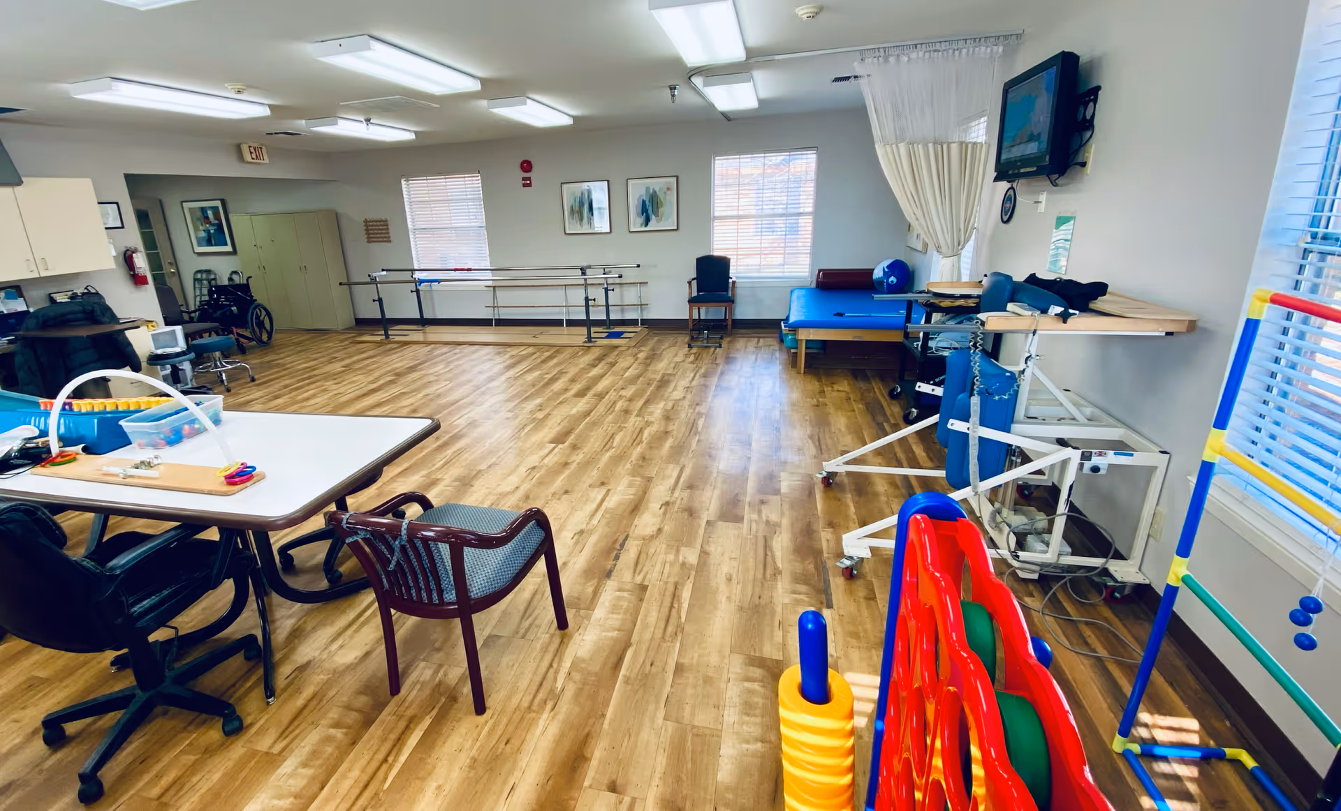A spacious therapy or activity room with wooden flooring, featuring a table with chairs, a wheelchair, parallel bars for walking exercises, a therapy bed, and various colorful therapy and recreational equipment. The room has windows with blinds, fluorescent ceiling lights, and a wall-mounted TV.