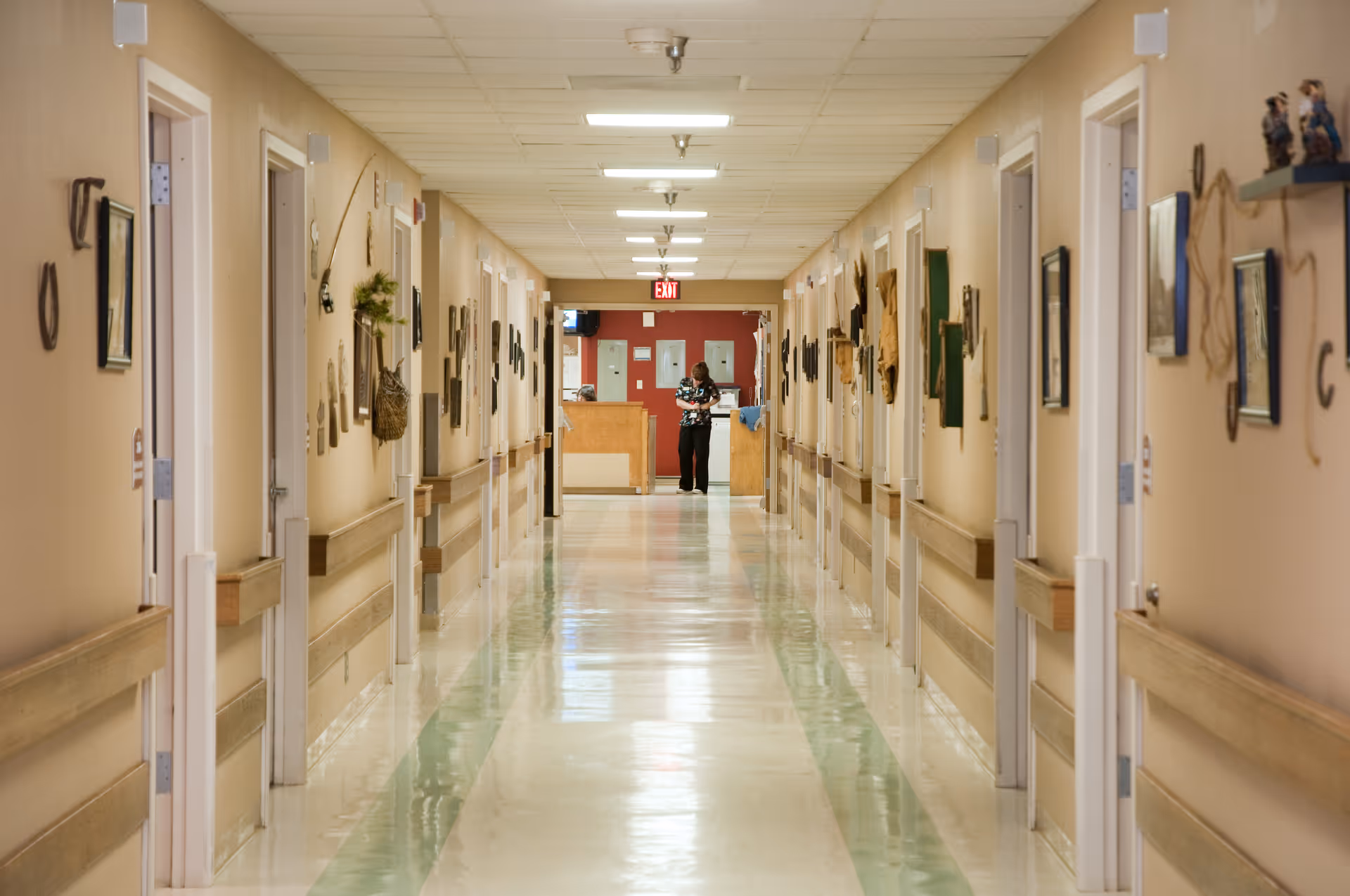 A long, clean hallway in a healthcare facility with beige walls and green and white tiled floor. Doors line both sides of the hallway, each decorated with various pictures and wall hangings. A person is standing at the far end near a reception or nurse station with a red wall behind them. The ceiling has fluorescent lights and handrails run along both sides of the hallway.
