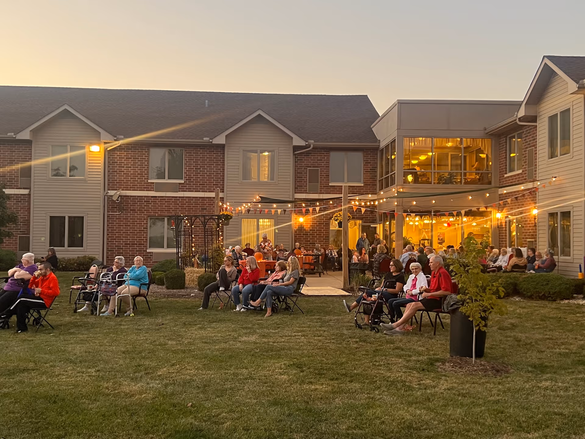 A group of elderly people sitting on chairs outside on a grassy area in front of a brick and siding building during dusk. String lights and small flags are hung above the gathering area, creating a warm and festive atmosphere. Some people are seated in pairs or small groups, enjoying the outdoor event.