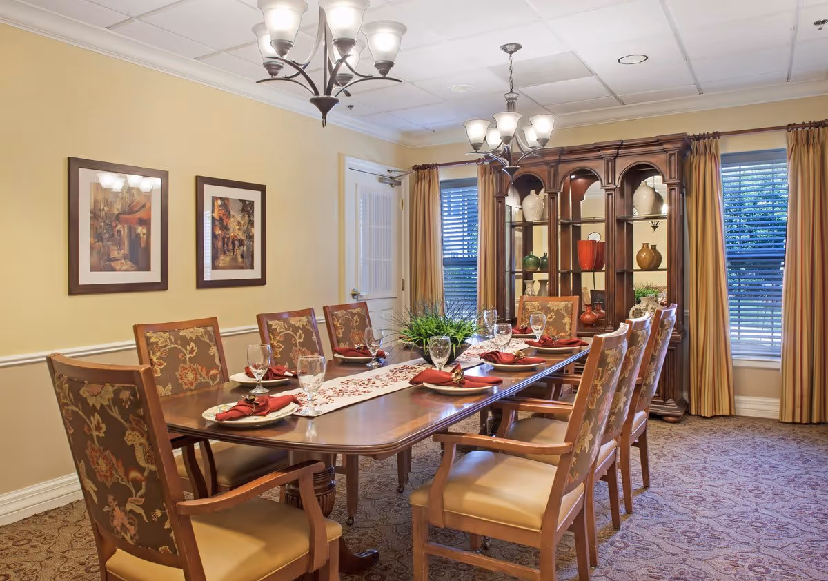 Formal dining room with a long table set for eight, upholstered chairs, chandeliers, and a wooden china cabinet by windows.