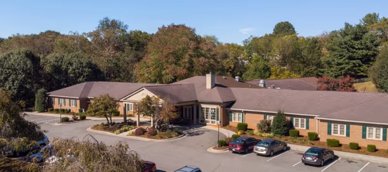 Exterior view of The Elms of Lynchburg senior living facility showing a single-story brick building with a covered entrance, surrounded by trees with autumn foliage and a parking lot with several cars.
