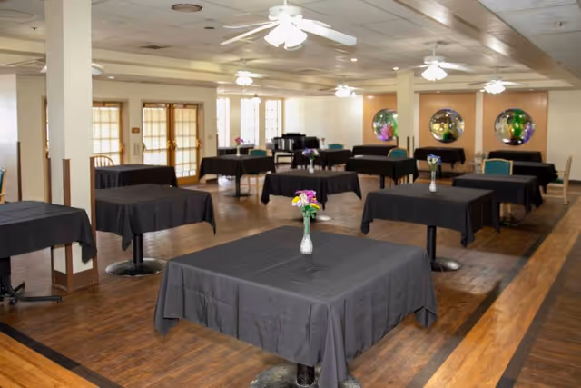 A spacious dining room with multiple square tables covered in black tablecloths, each table adorned with a small vase of colorful flowers. The room has wooden floors, ceiling fans with lights, and round mirrors on one wall. Large windows and glass doors allow natural light to enter the space.