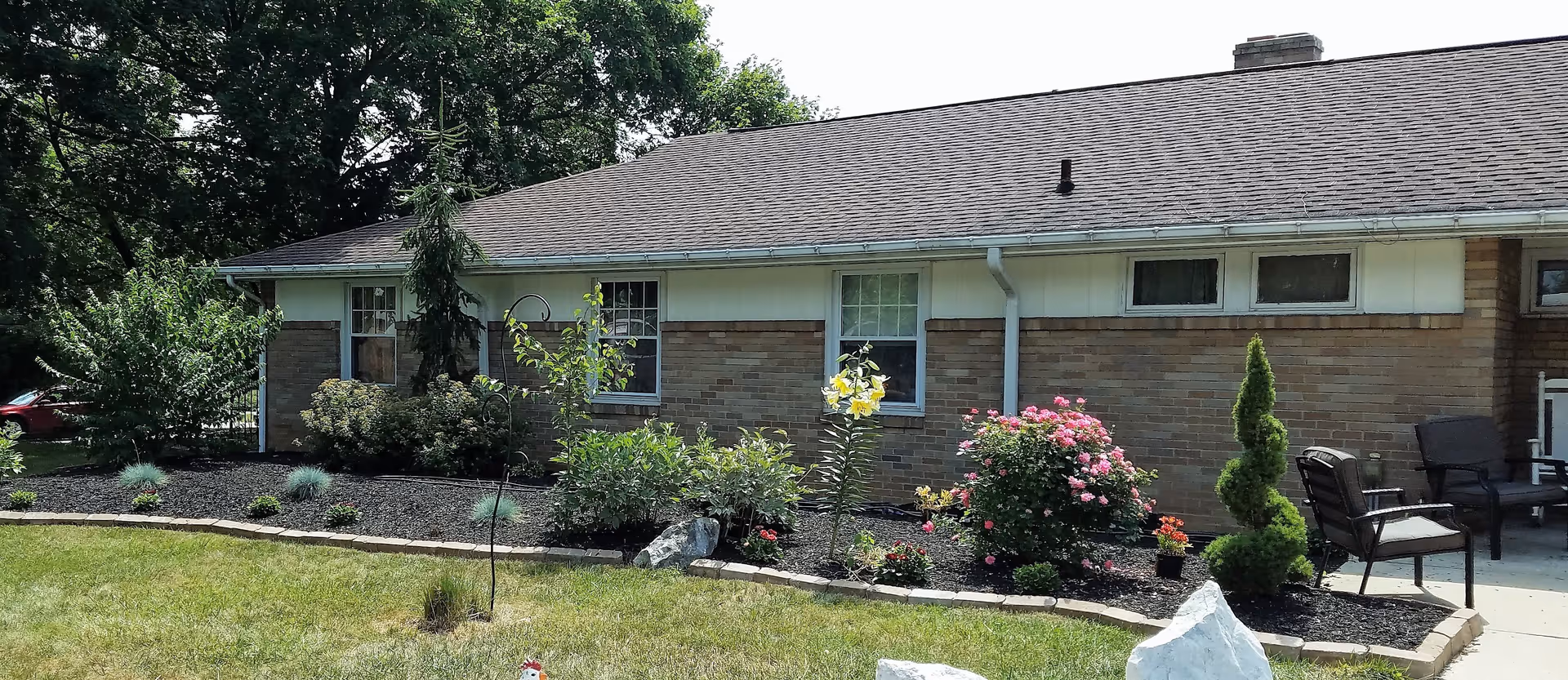 Exterior view of a single-story brick building with a sloped roof, surrounded by a well-maintained garden featuring various shrubs, flowering plants, and small trees. There are two windows visible on the building and outdoor seating with chairs on a concrete patio area to the right.