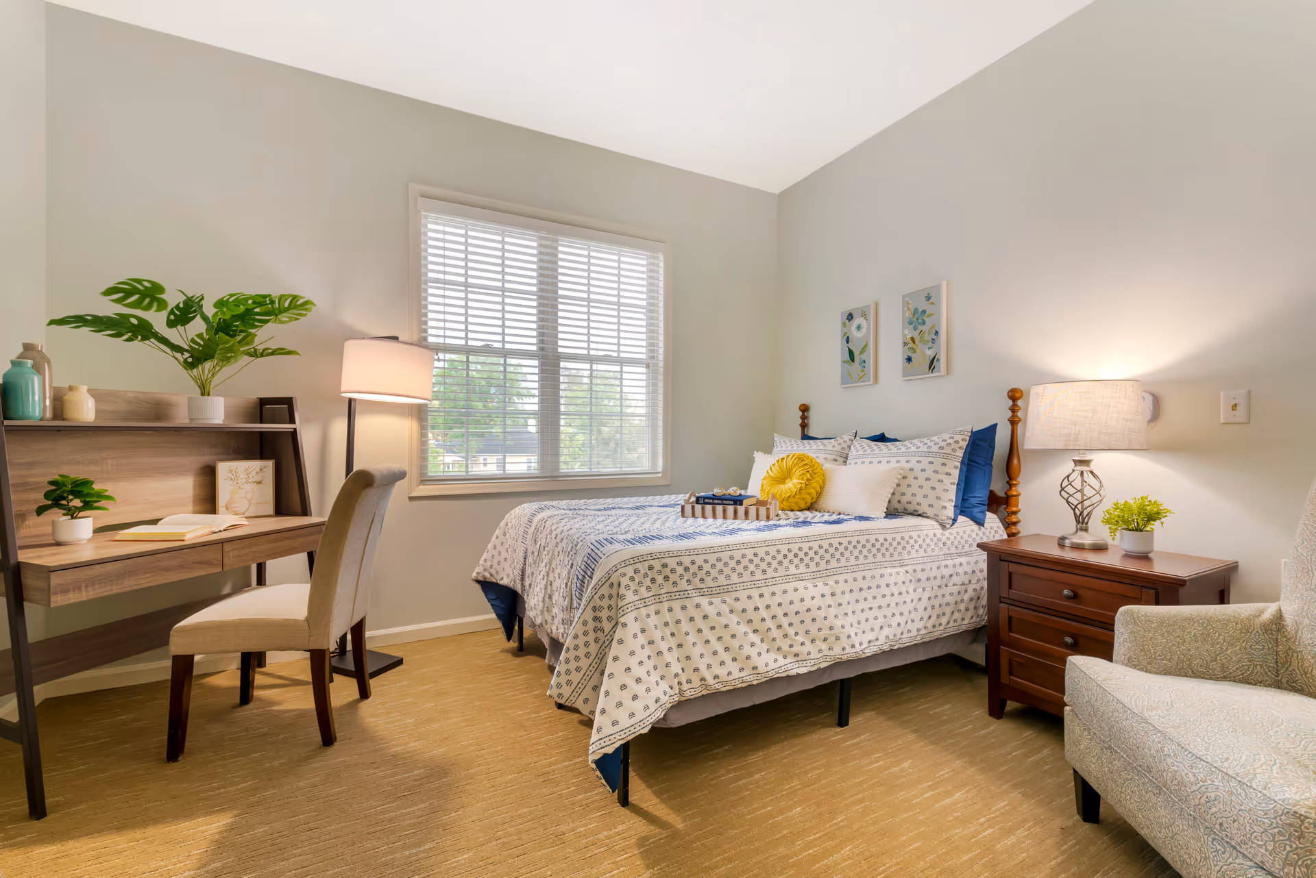 Sunlit furnished bedroom with a bed, bedside table and lamp, a desk with chair, and an armchair.