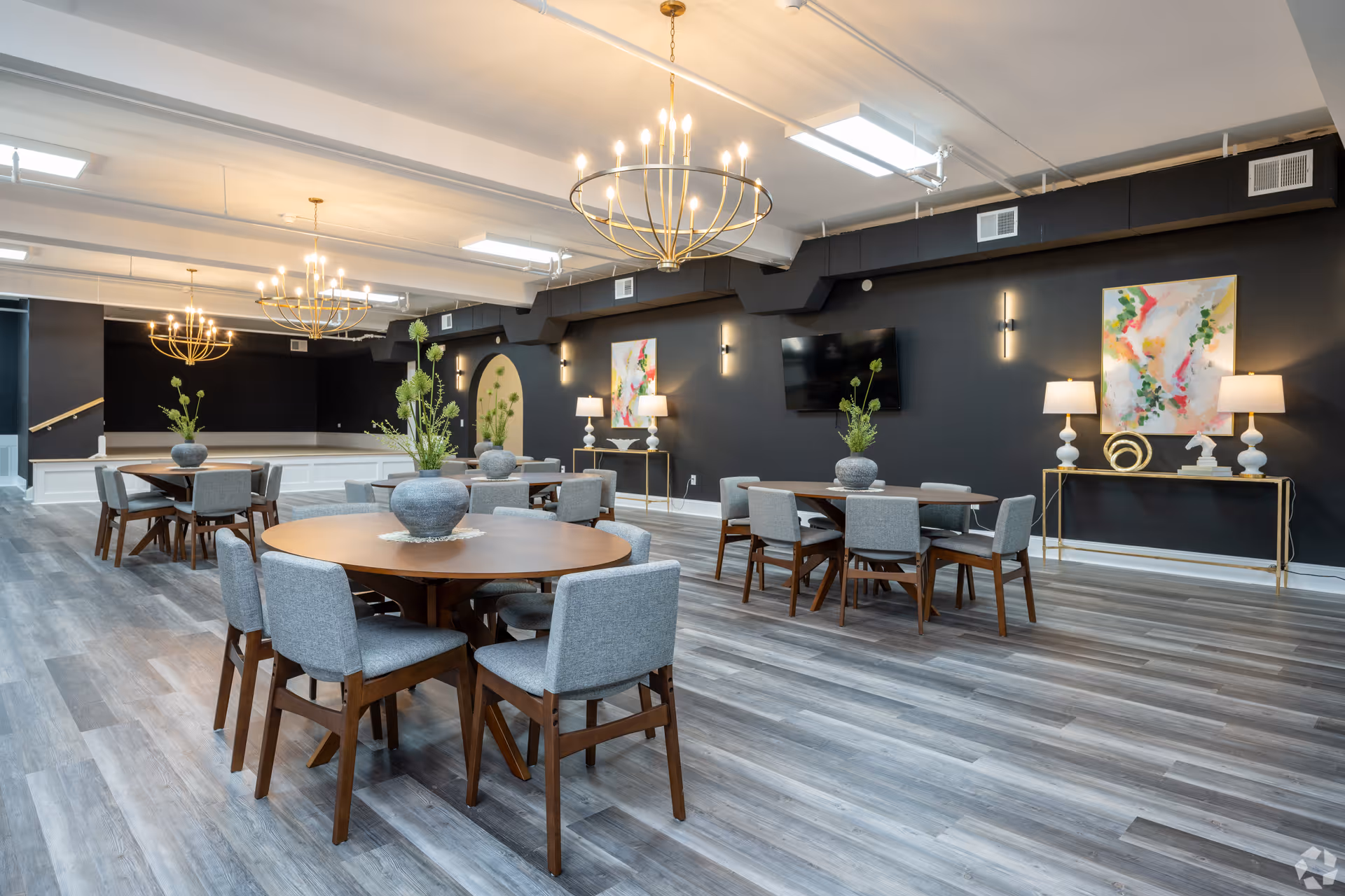 A spacious, modern dining room with round and rectangular wooden tables surrounded by gray upholstered chairs. The room features light gray wood flooring, dark gray walls, contemporary gold chandeliers, and decorative plants on the tables. There are two abstract paintings on the walls, a flat-screen TV, and console tables with lamps and decorative items.
