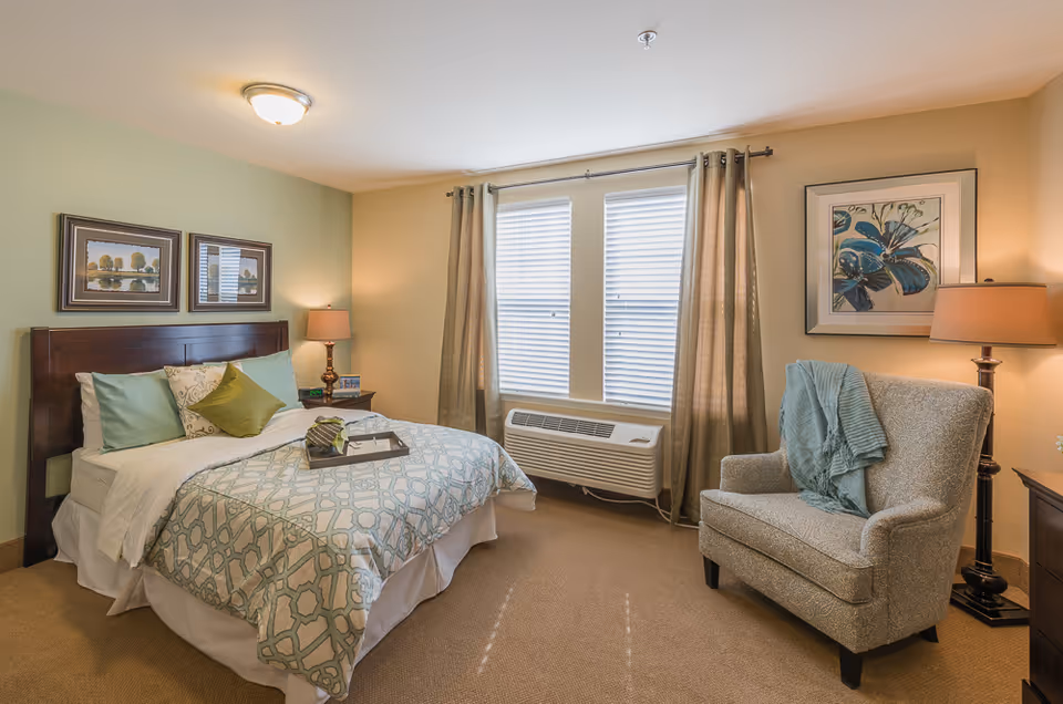 A cozy bedroom with a neatly made bed featuring patterned bedding and multiple pillows. There are two framed pictures above the wooden headboard. A bedside table with a lamp and some items is next to the bed. Across from the bed is a comfortable upholstered armchair with a blue throw draped over it. The room has two windows with blinds and beige curtains, and a wall-mounted air conditioning unit below the windows. The walls are painted in soft green and beige tones, and there is a framed floral artwork on the wall near the armchair.
