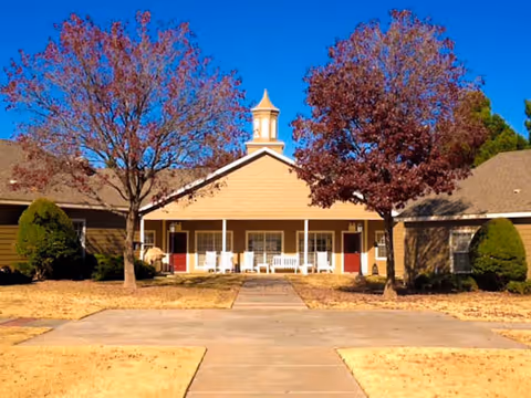 Front exterior view of a single-story building with a central cupola, flanked by two trees with reddish leaves and trimmed bushes on either side. The building has a covered porch with white rocking chairs and red doors.
