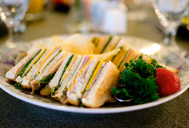 A plate with several triangular club sandwiches filled with layers of turkey, cheese, lettuce, and tomato, accompanied by a sprig of parsley, a strawberry, and potato chips on the side.