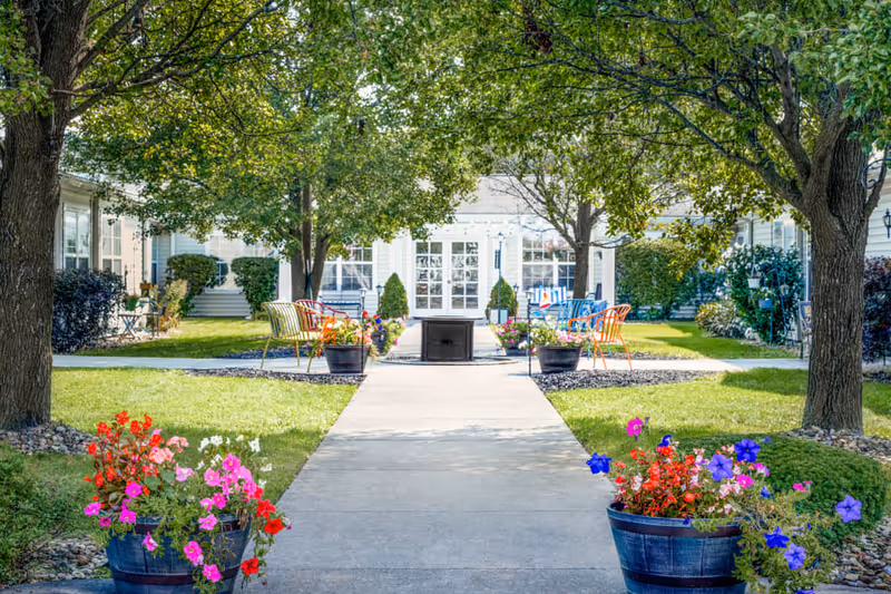A peaceful outdoor courtyard with a concrete walkway flanked by colorful flower pots and green grass. There are trees providing shade, and several colorful chairs arranged around a central fire pit. The background shows a white building with large windows and doors.
