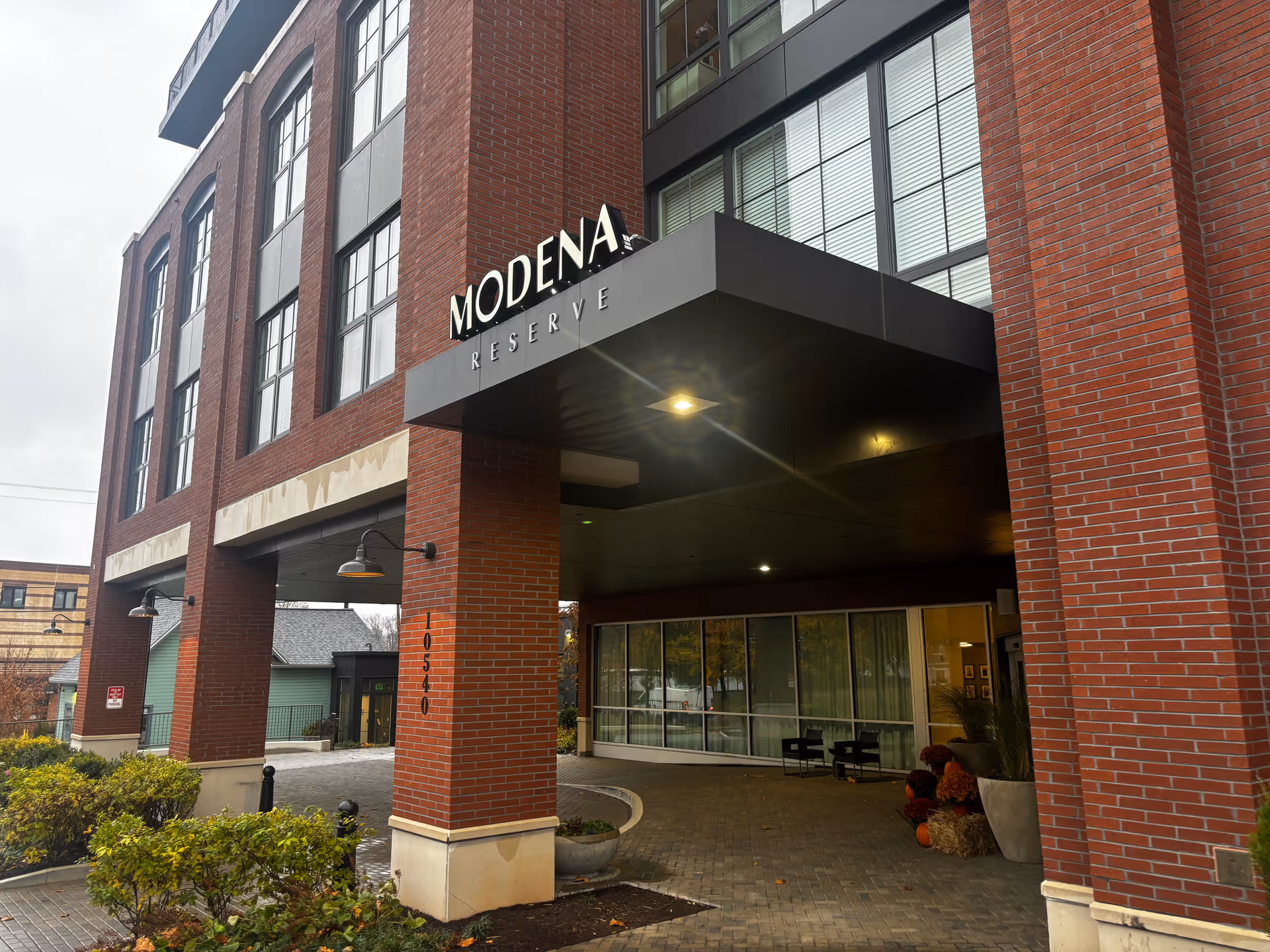 Entrance of a multi-story brick building with large windows and a covered driveway. The building has a sign that reads 'MODENA RESERVE'. There are some plants and bushes near the entrance and a couple of chairs and decorations under the covered area.
