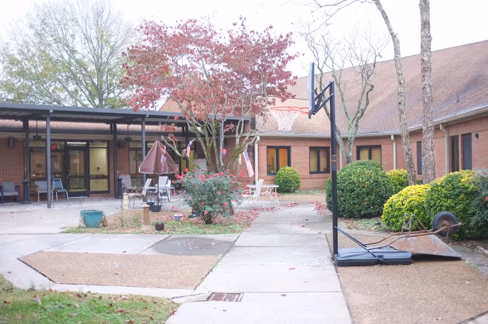 Outdoor courtyard area of Knowles Home Assisted Living with a basketball hoop, patio chairs, a table with an umbrella, bushes, a tree with red leaves, and a brick building surrounding the courtyard.