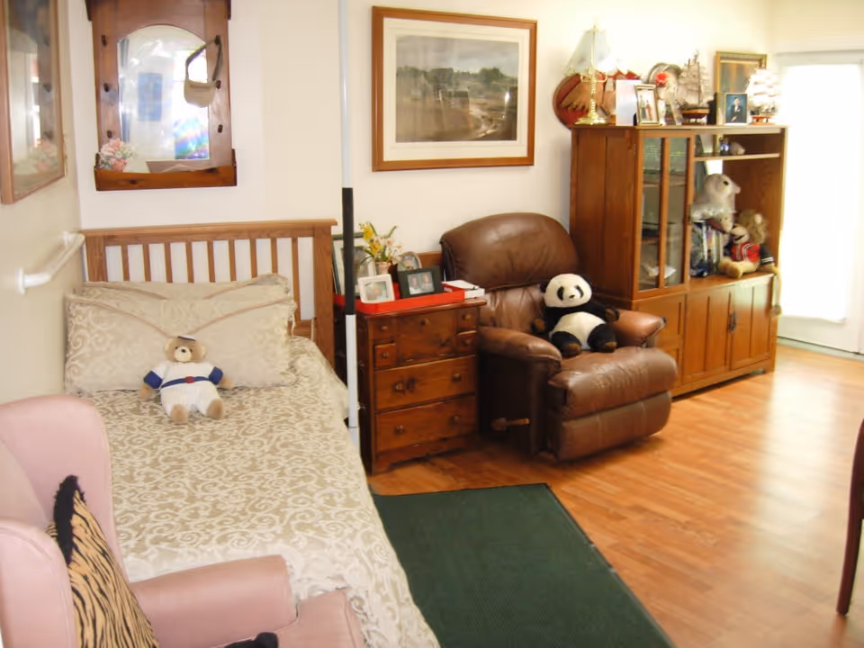 A cozy room with a single bed covered in a patterned beige bedspread and a teddy bear dressed in a baseball uniform sitting on it. Next to the bed is a wooden nightstand with framed photos and flowers. A brown leather recliner chair with a stuffed panda sits beside the nightstand. A wooden cabinet with glass doors displays various decorative items and stuffed animals. The room has wooden flooring and a green rug, with framed artwork on the walls and a window letting in natural light.