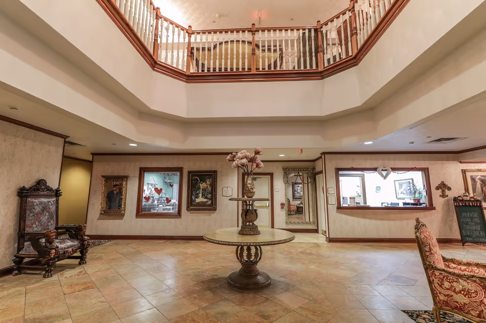 Spacious lobby area with a round table in the center holding a decorative vase with flowers. The room features tiled flooring, ornate chairs with patterned upholstery, framed artwork on the walls, and a balcony railing above. There is a reception window on the right side and a large mirror on the back wall.