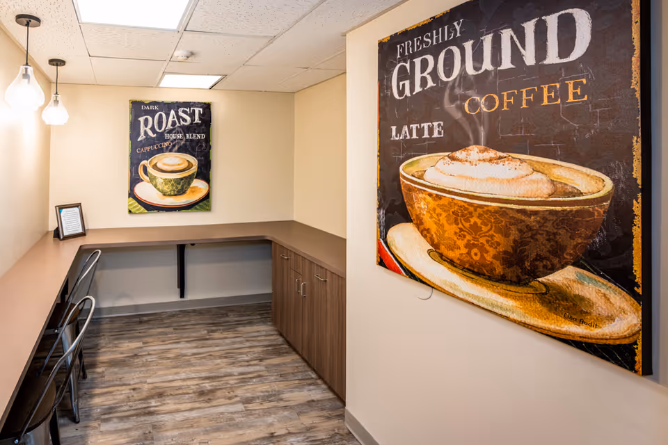 A small coffee nook with a long brown countertop and two metal chairs. The walls are decorated with two large coffee-themed paintings, one showing a cup of cappuccino with the words 'Dark Roast House Blend' and the other showing a latte with the words 'Freshly Ground Coffee Latte'. The floor has wood-like vinyl flooring and there are two pendant lights hanging from the ceiling.