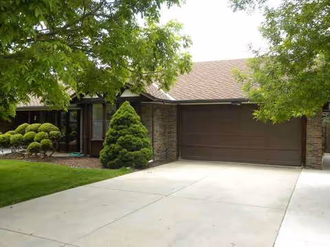 A single-story assisted living residence with a brown garage door, driveway, and manicured greenery.