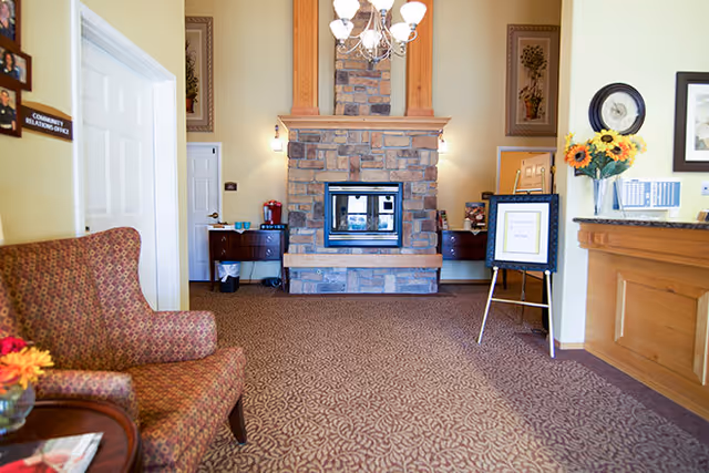 Interior view of a senior living facility lobby with a patterned carpet, a stone fireplace in the center, a chandelier above, a cushioned armchair on the left, a wooden reception desk on the right with a vase of sunflowers, and a sign on an easel near the fireplace.