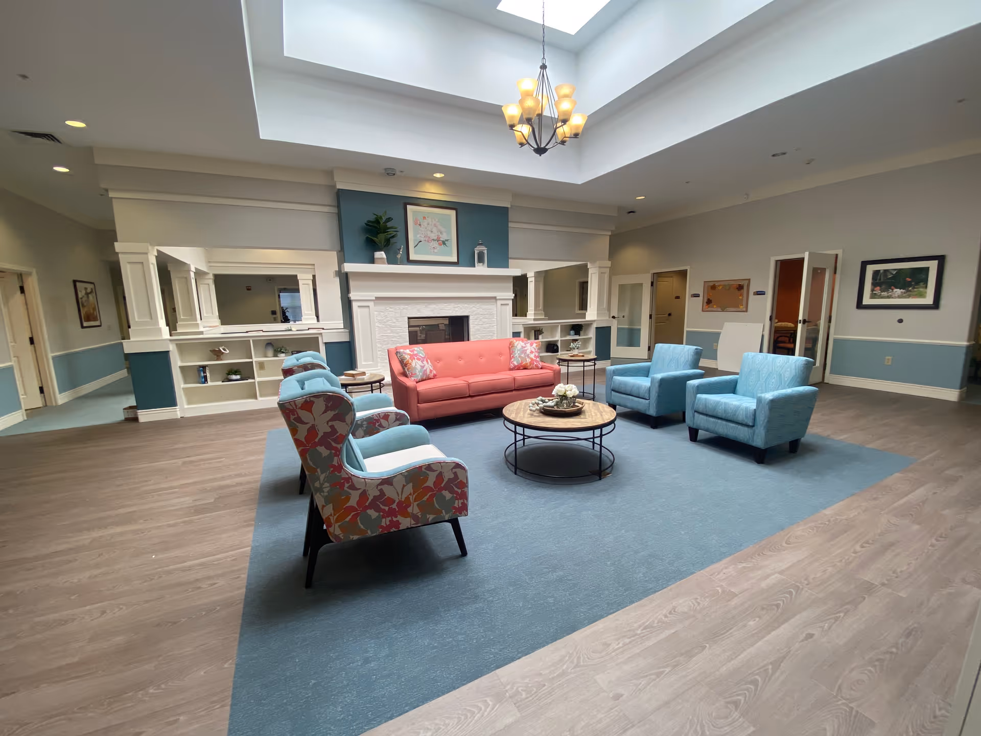 A spacious and well-lit living room area with a large skylight overhead. The room features a coral pink sofa, two light blue armchairs, and two floral patterned chairs arranged around a round wooden coffee table on a blue area rug. The walls are painted in light blue and white, with built-in shelves and a white fireplace in the center. There are framed pictures and decorative items on the walls and shelves.