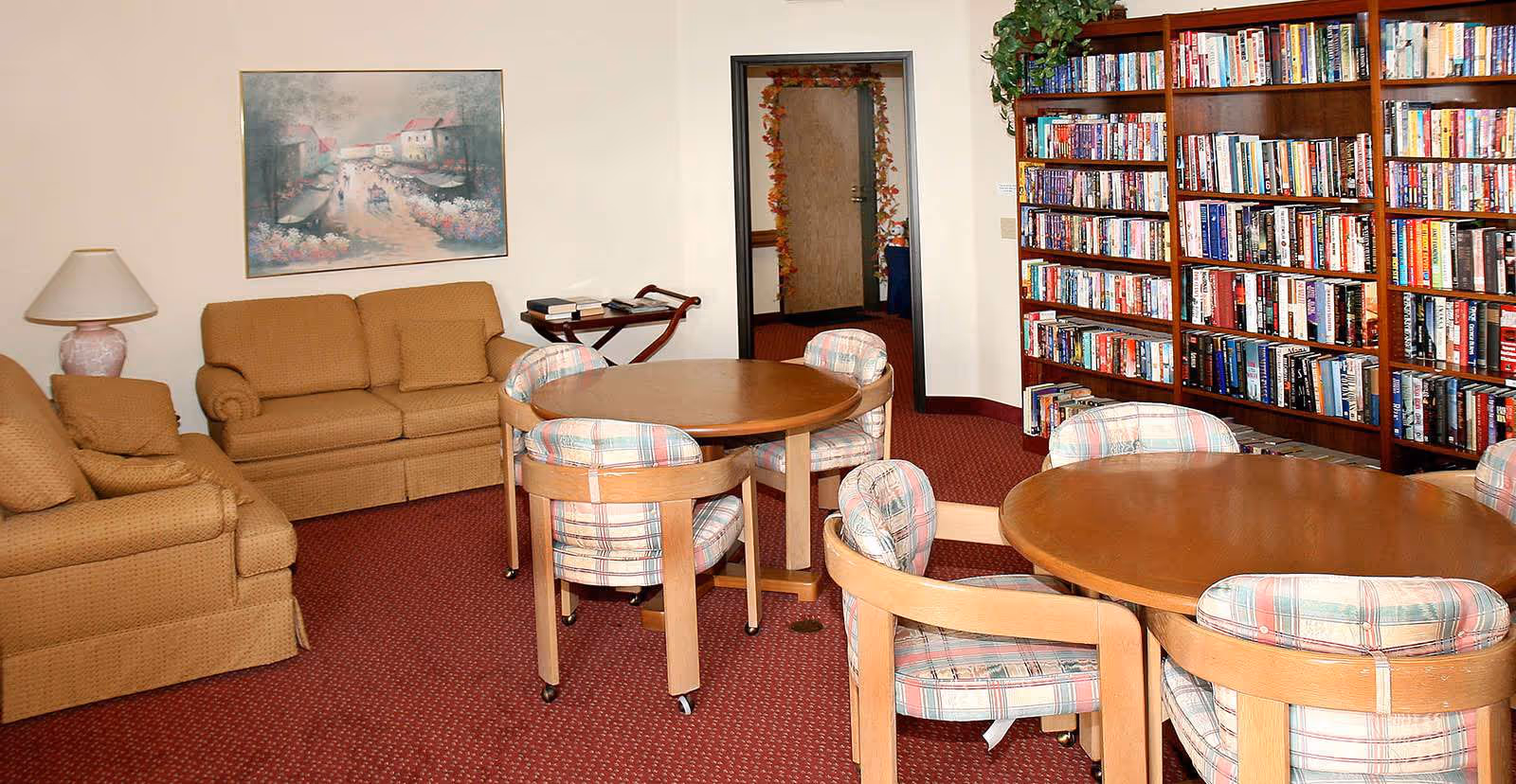 Carpeted common room with sofas, round wooden tables and cushioned chairs facing a large bookshelf filled with books.