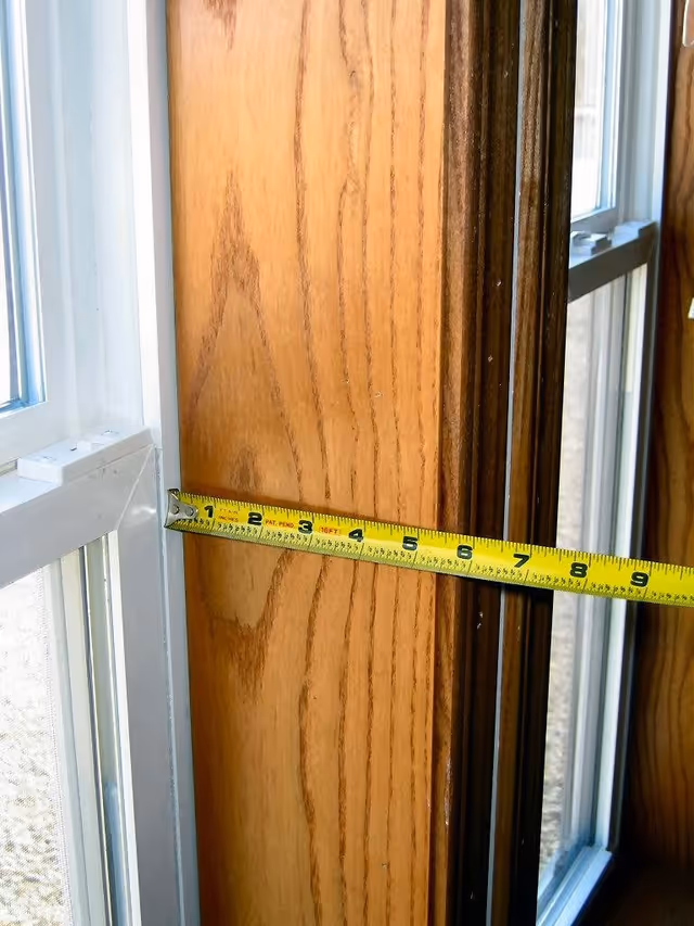 A close-up view of a wooden window frame with a yellow measuring tape stretched across it, showing a measurement of about 8 inches. The window has white trim and glass panes, with natural light coming through.