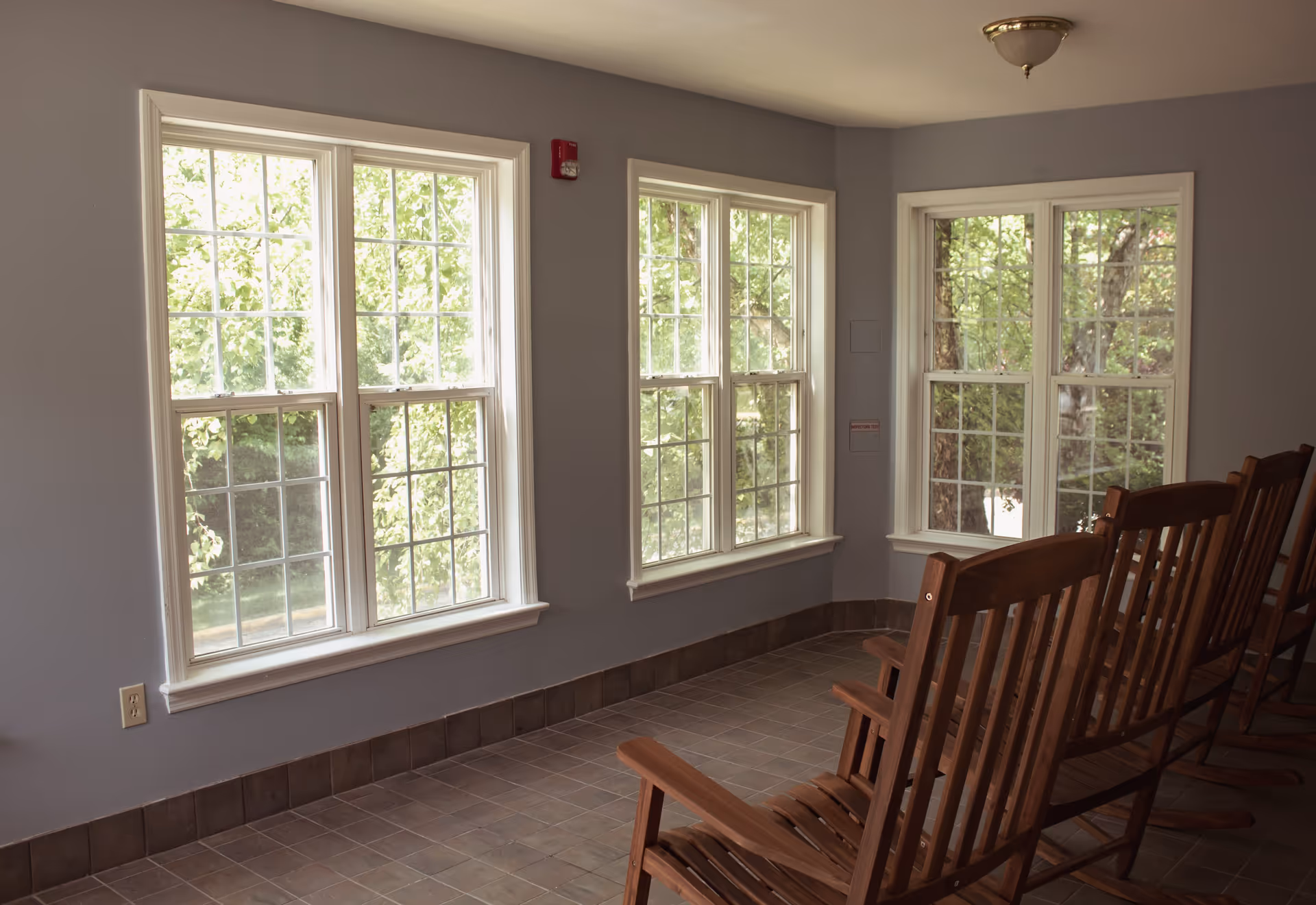 A quiet interior corner of a room with three large windows showing green trees outside. Four wooden rocking chairs are lined up facing the windows. The walls are painted light gray and the floor is tiled with brown tiles.