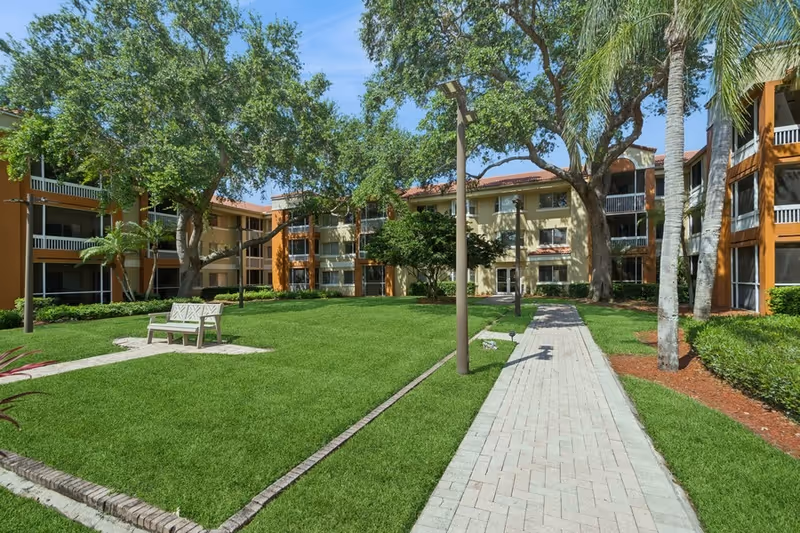 Outdoor courtyard area of a senior living facility with well-maintained green grass, a paved walkway, benches, tall trees, and a multi-story building with balconies surrounding the courtyard under a clear blue sky.