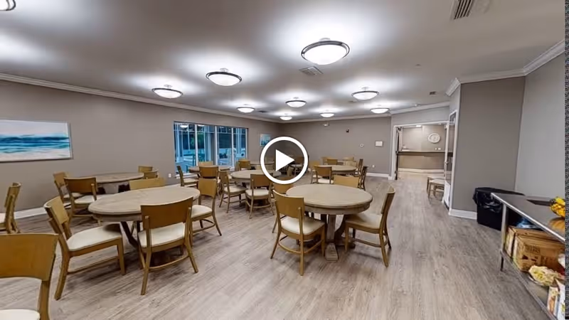 A spacious dining room with multiple round wooden tables and chairs arranged neatly. The room has light-colored wooden flooring, beige walls, and several ceiling lights. There is a large window on one side and a serving area or counter visible in the background.