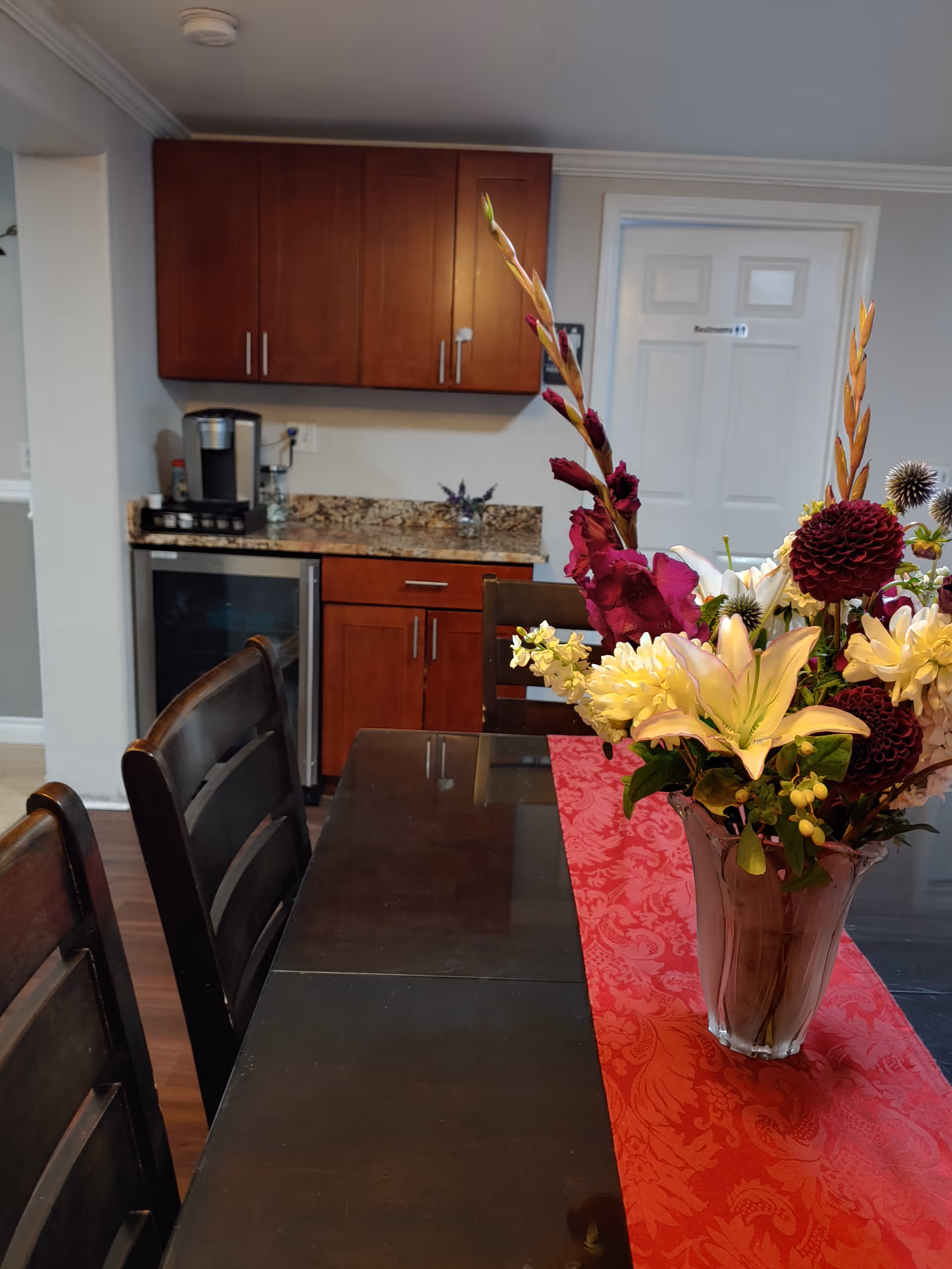 Dining table with a red runner and vase of flowers in a room with wooden chairs, a small kitchenette and a door labeled 'Restrooms'.