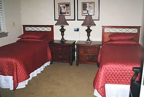 Twin beds with matching red quilts, nightstands, lamps, and framed pictures in a beige carpeted bedroom.