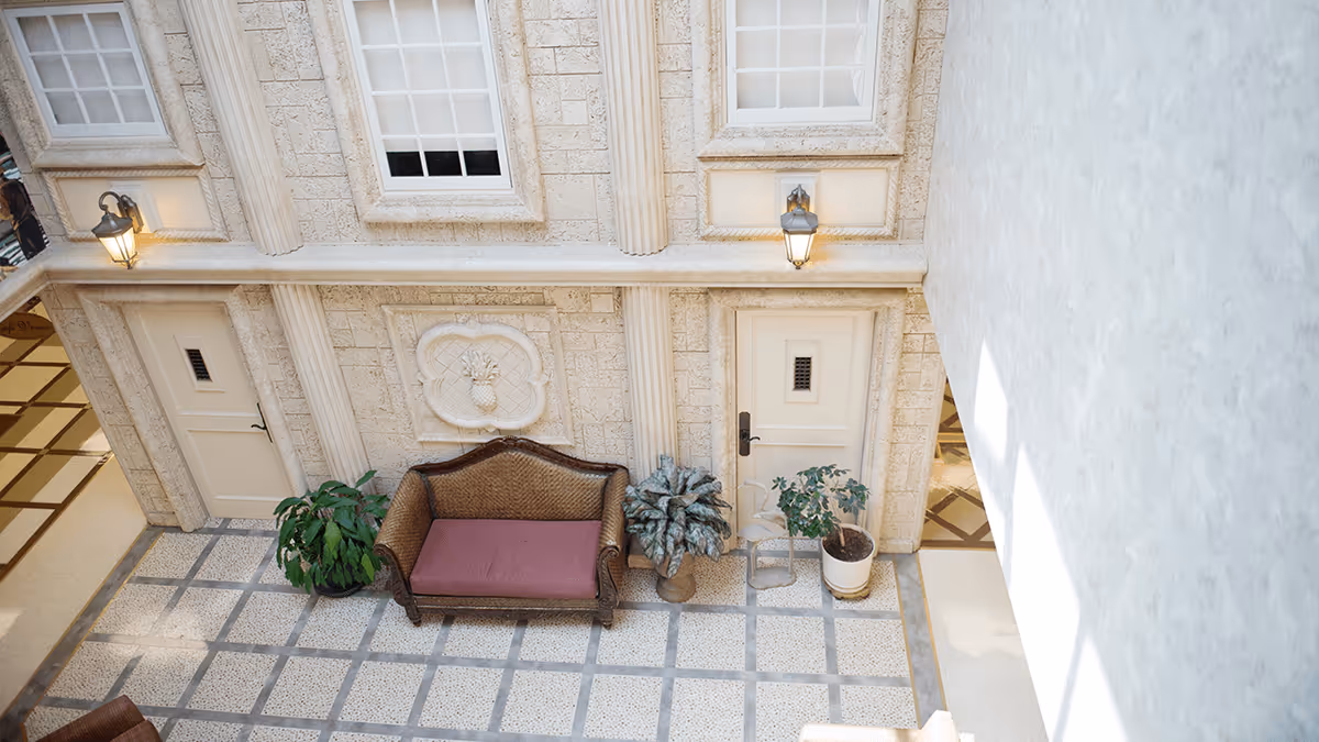 Indoor courtyard-style hallway with a decorative stone wall, two doors, a wicker bench with a pink cushion, potted plants, wall lanterns, and a tiled floor.