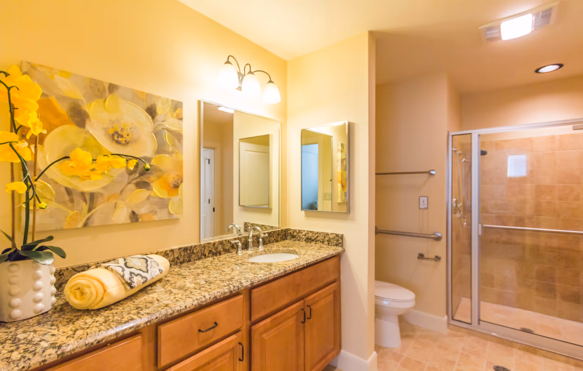 Well-lit bathroom featuring a granite vanity with sink and mirror, decorative yellow flowers, a toilet, and a glass-enclosed tiled shower.