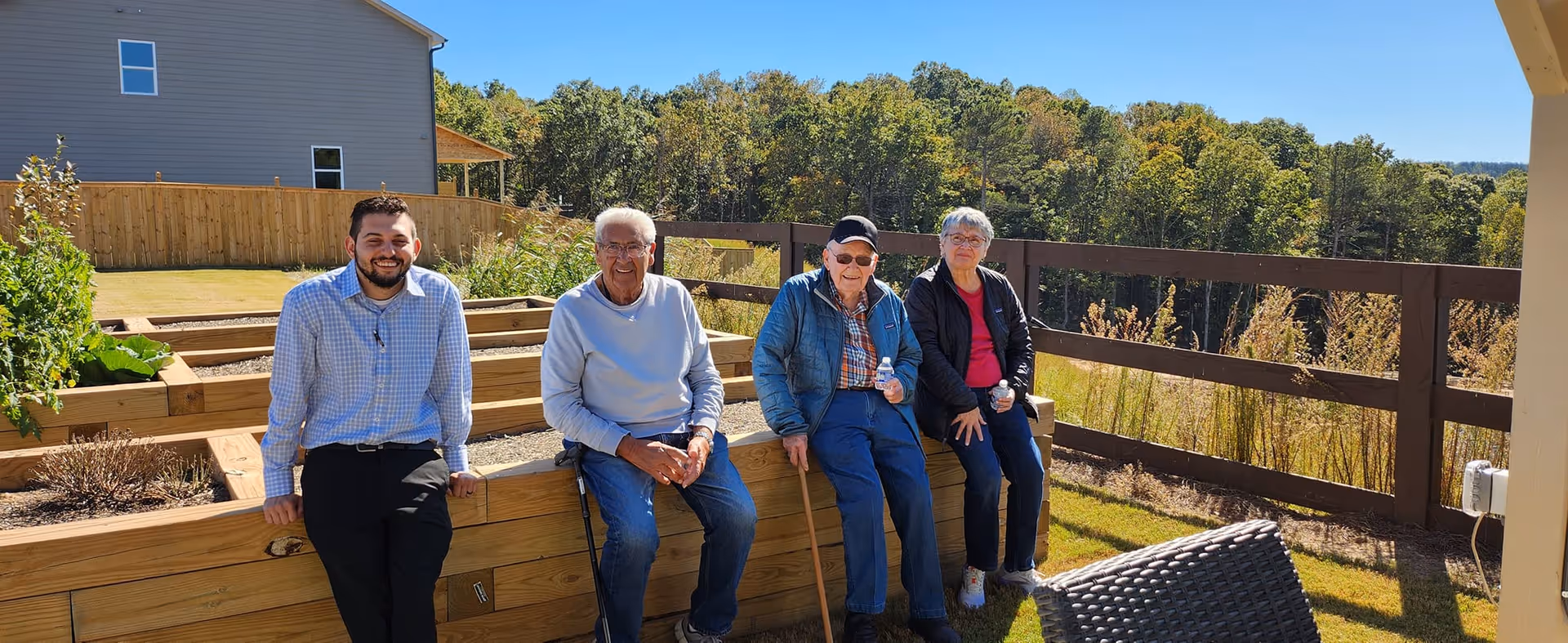 Four people sitting outdoors on a wooden raised garden bed structure. Three elderly individuals and one younger man are smiling and enjoying a sunny day with trees and a house in the background.