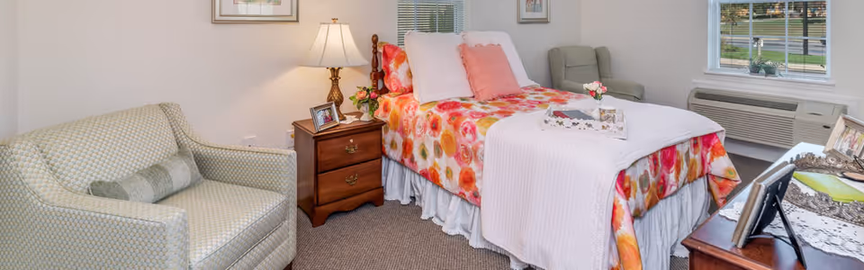 A bright, cozy bedroom with a floral-covered single bed, bedside table with lamp, armchair, and a window with an air conditioner below it.