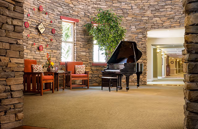 Stone-walled common lounge featuring a grand piano, red upholstered chairs, and a decorative wall clock.
