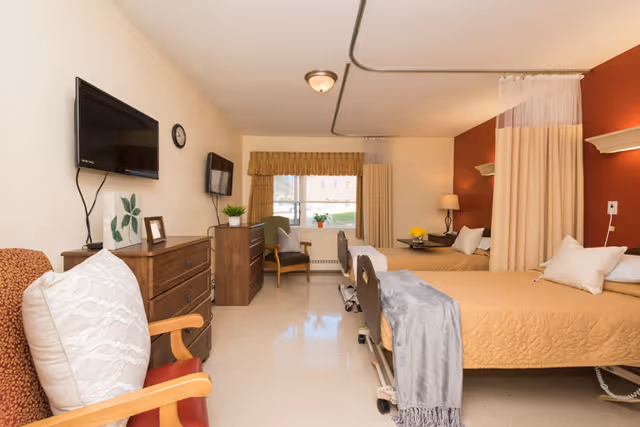 Shared nursing facility bedroom with two hospital-style beds, dressers, chairs, a wall-mounted TV, window, and a privacy curtain.
