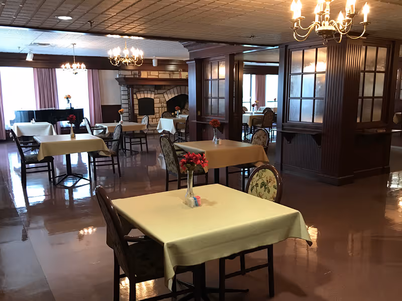 Dining room with multiple tables draped in tablecloths and floral centerpieces, chandeliers overhead, and a stone fireplace and piano in the background.