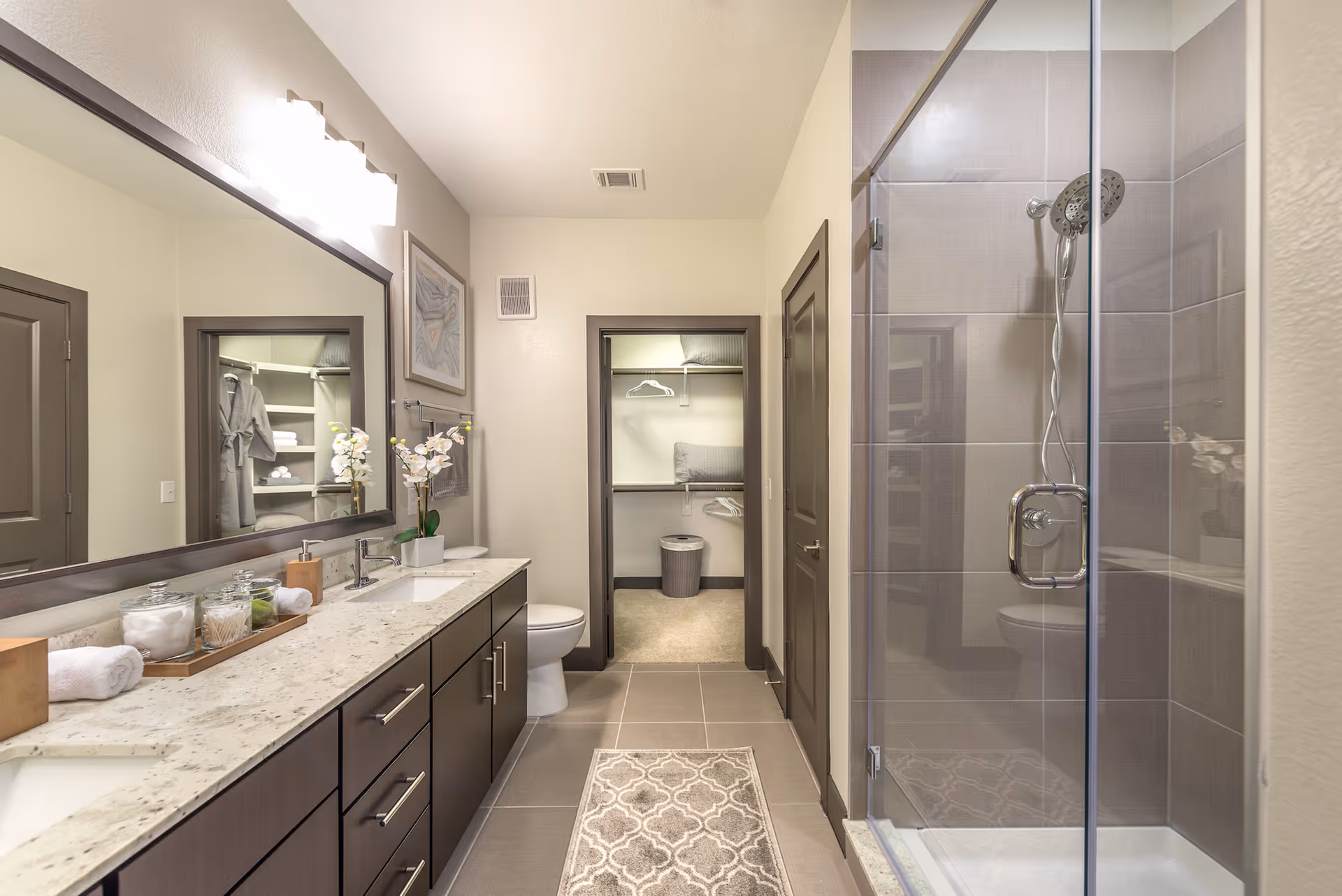 A modern bathroom with a double sink vanity featuring a granite countertop and dark wood cabinets. There is a large mirror above the sinks, a glass-enclosed shower with a chrome showerhead, a toilet, and a doorway leading to a walk-in closet with shelves and hanging space. The bathroom has neutral-colored tiles and walls, with a patterned rug on the floor and decorative items like a potted orchid and jars on the countertop.
