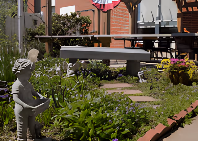 A garden area with green plants and purple flowers, featuring a small statue of a child holding a bowl. There is a stone bench and a picnic table in the background, along with a brick wall and some outdoor decorations.