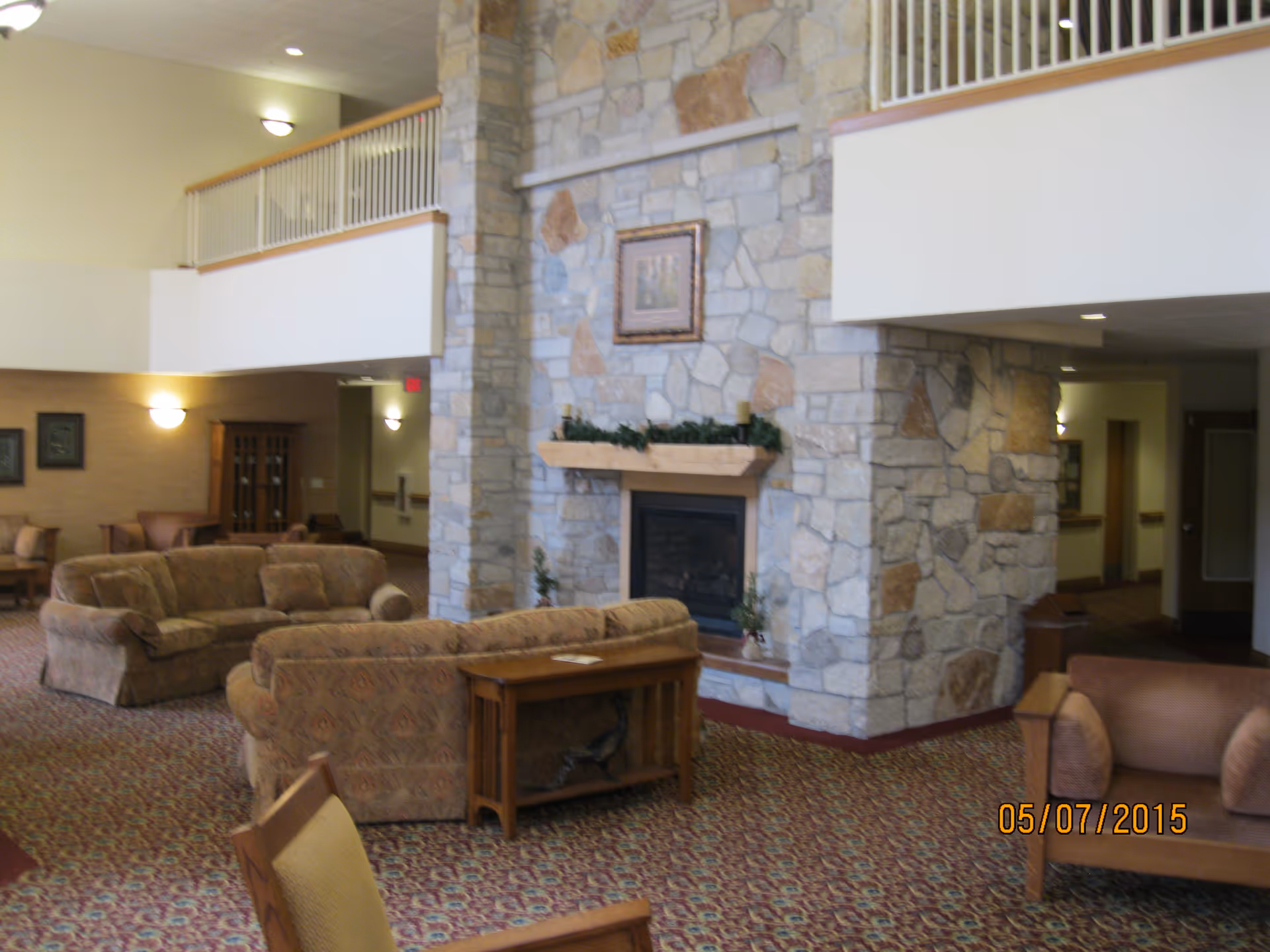 Spacious assisted living facility common area with patterned carpet, multiple upholstered sofas and chairs arranged around a large stone fireplace. The room has high ceilings with a balcony railing overlooking the area and soft wall lighting.
