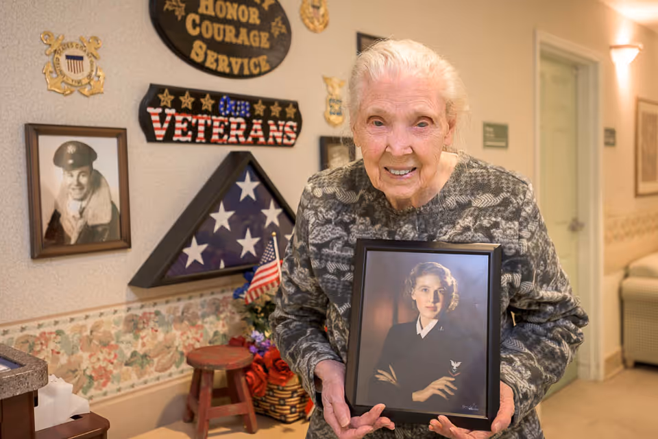 An elderly woman with white hair and a patterned sweater is standing in a hallway holding a framed portrait of a younger woman in a military uniform. Behind her, the wall is decorated with a folded American flag in a triangular display case, a sign that reads 'Honor Courage Service Our Veterans,' and a black and white photo of a man in military attire. There is a small stool and some flowers beneath the display.