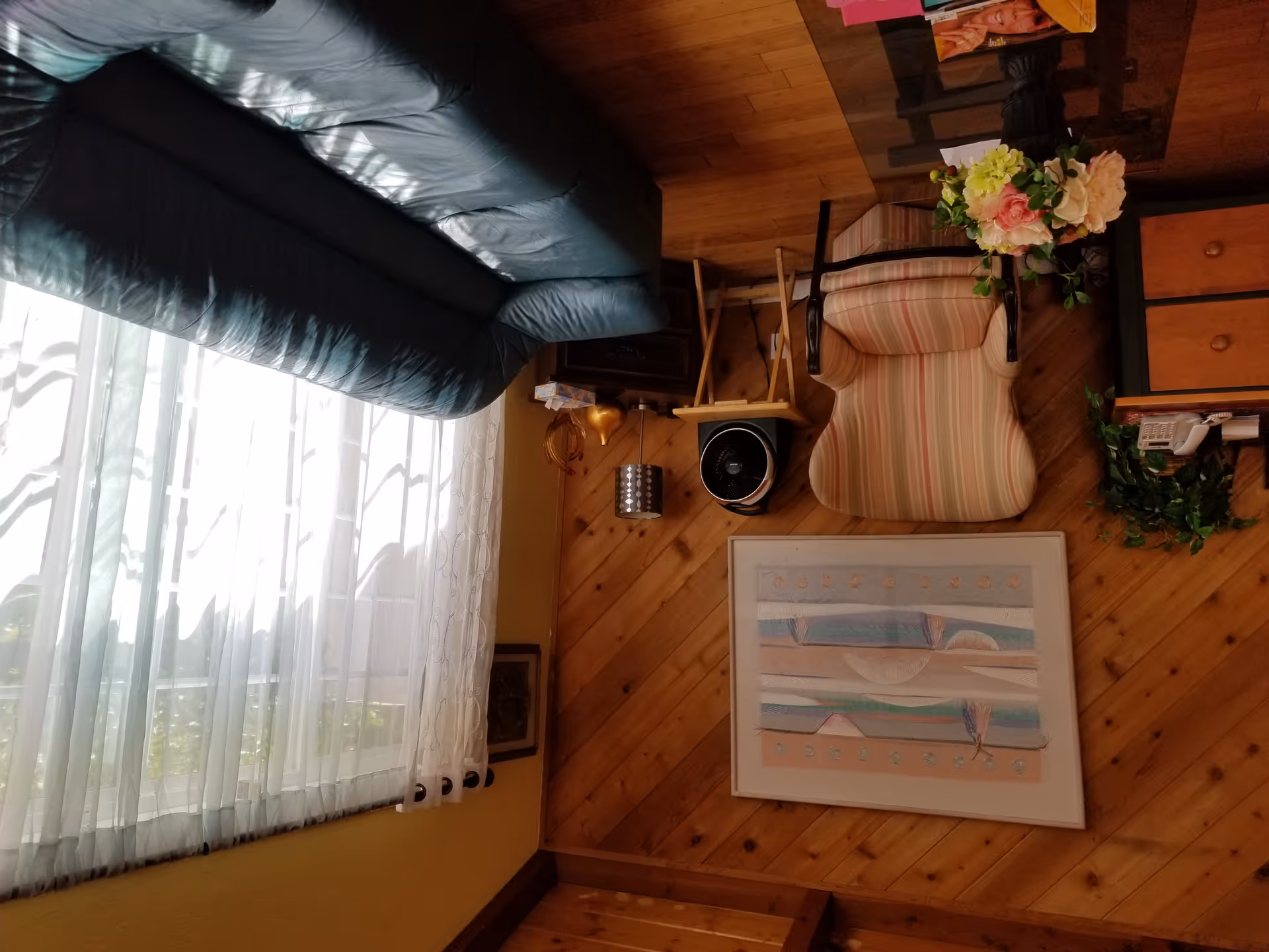 A cozy living room with wooden paneled walls, a striped armchair, a dark green leather sofa, a glass coffee table with a flower arrangement, a small wooden side table with a fan and lamp, and sheer white curtains covering a large window.